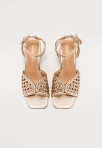 Sandales à talons dorés avec une tige tressée, une pointe carrée et une bride à la cheville. Fini métallique lisse sur le talon et la semelle intérieure.