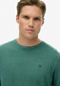 Camiseta de algodón verde con cuello redondo, que presenta un sutil logo bordado en el lado izquierdo del pecho, textura suave y mangas cortas.