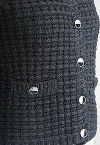 Gebreide donkergrijze vest met een gestructureerd patroon. Voorzien van zilverkleurige knopen en een voorzak met bijpassende knop.