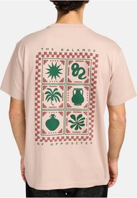 Lichtroze T-shirt met een centrale afbeelding van groene abstracte vormen, omrand door roodachtige vierkanten. De tekst luidt: "DE BALANS VAN TEGENSPOEDEN."