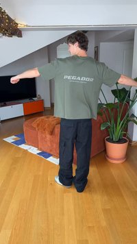 Olive green short-sleeve T-shirt with “PEGADOR” printed on the back, paired with black cargo pants in a casual indoor setting.