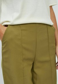 Pantalons olive en tissu lisse, avec une coupe ajustée, des poches latérales et des détails de couture à l'avant.