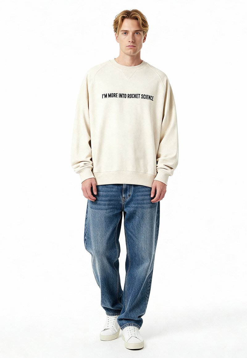 Beige sweatshirt met zwarte tekst "I'M MORE INTO ROCKET SCIENCE," ronde hals, raglanmouwen, gecombineerd met lichtblauwe jeans en witte sneakers.