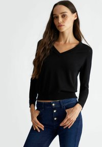 Pullover nero a maglia con scollatura a V e maniche lunghe, con polsini e orlo a coste. Abbinato a jeans in denim blu a vita alta.