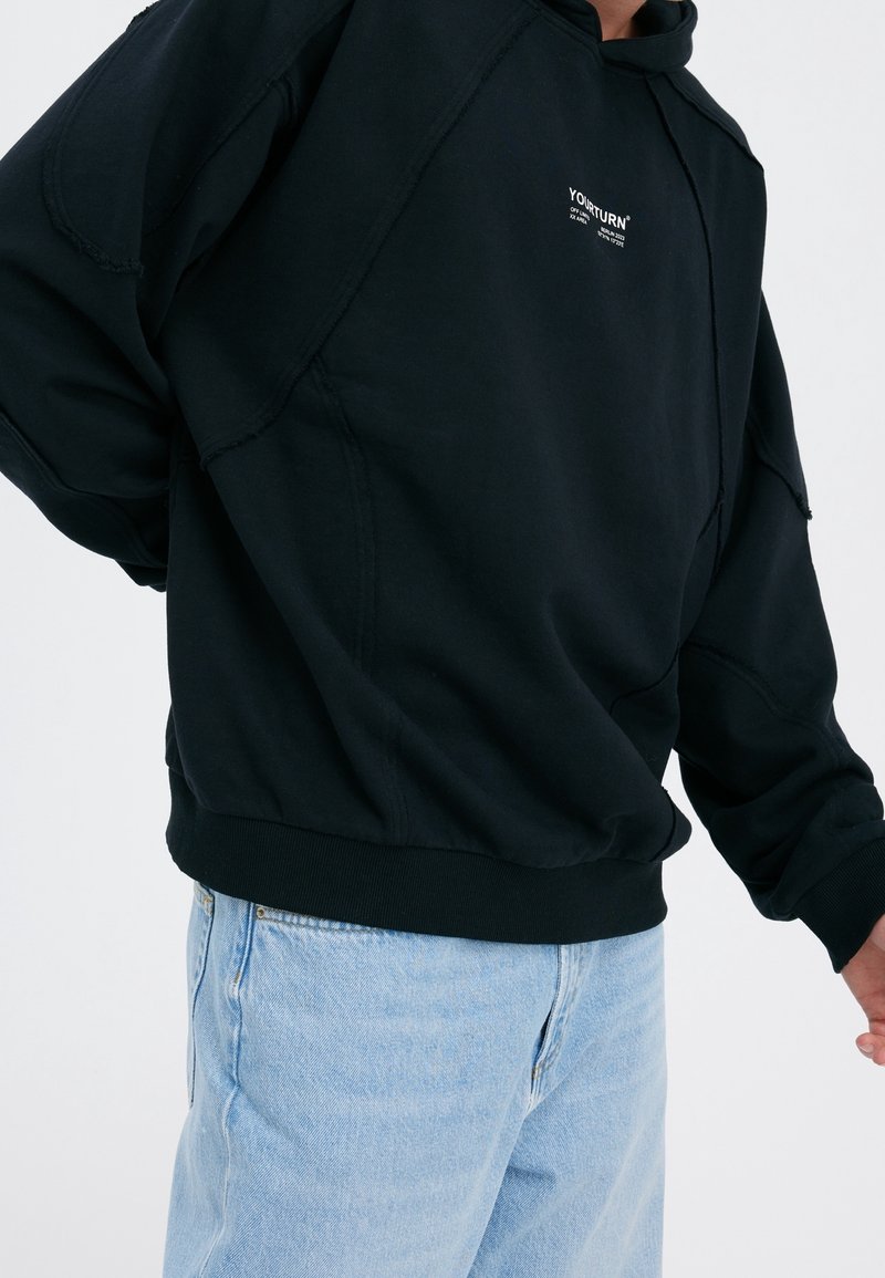 Sudadera negra de cuello redondo con un ajuste relajado, detalles cosidos y un pequeño logo blanco en el pecho, combinada con jeans azul claro.