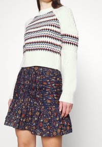 Femme portant un pull en maille blanc à motifs avec des rayures rouges, bleues et jaunes, associé à une mini-jupe à volants bleu marine à motifs floraux.