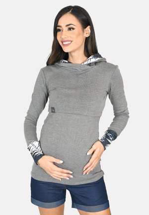 Lächende schwangere Frau mit langen dunklen Haaren hält ihren Bauch, trägt einen grauen Hoodie mit gemusterten Bündchen und marineblaue, hochgekrempelte Shorts.