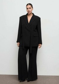 Czarny garnitur szyty na miarę z oversize'ową marynarką i spodniami o szerokich nogawkach. Charakterystyczne cechy to ostre klapy, zarysowana sylwetka oraz minimalistyczny design.