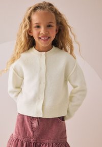 Ivori krāsas adīts kardigans ar apaļu kaklu un pogu aizdari, kopā ar rozeādas kordurojas svārkiem ar kabatām un volāniem apakšmalā.