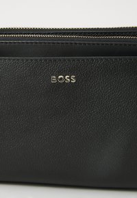 Zwarte leren tas met een gestructureerde afwerking, voorzien van een gouden "BOSS"-logo en dubbele ritssluitingen aan de bovenkant voor veilige opberging.
