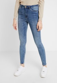 Jean bleu slim porté avec un pull beige et des baskets blanches, montrant la partie médiane jusqu'aux pieds sur fond blanc.