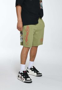 Pantaloni corti verde oliva con testo grafico rosso, caratterizzati da una stampa dettagliata sul lato. Abbinati a scarpe da ginnastica bianco e nere e calzini bianchi.