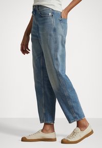 Ljusblå denimjeans med avslappnad passform, blekt utseende och slitna detaljer. Står i kombination med beige avslappnade sneakers.