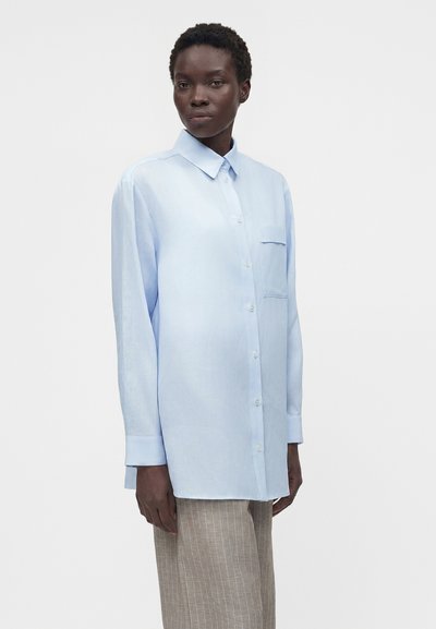 Γυναίκα με κοντά σγουρά μαλλιά φορώντας ένα ανοιχτό μπλε πουκάμισο μεγάλης διάστασης με κουμπιά και μπεζ παντελόνι με ριγέ σχέδιο σε απλό φόντο.