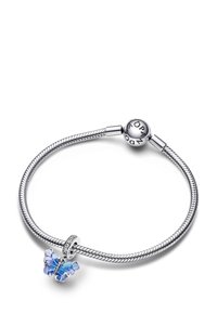 Strieborný náramok s čarom so spletitým dizajnom, obsahujúci charm vo forme modrého motýľa s priehľadnými krídlami a kryštálovými akcentmi.