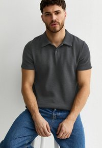 Joven con cabello rizado corto, vestido con una camiseta tipo polo de manga corta gris oscuro y jeans azules, sentado contra una pared blanca lisa.
