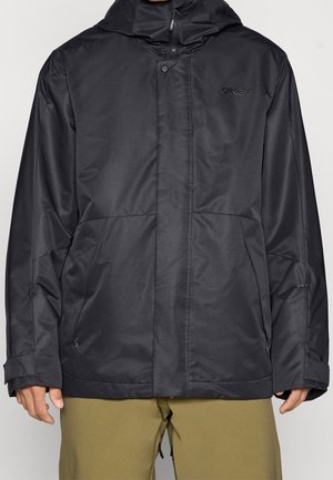 Veste à capuche noire Oakley avec fermeture éclair frontale et bouton-pression, portée par une personne avec un pantalon vert olive, mains visibles sur les côtés.