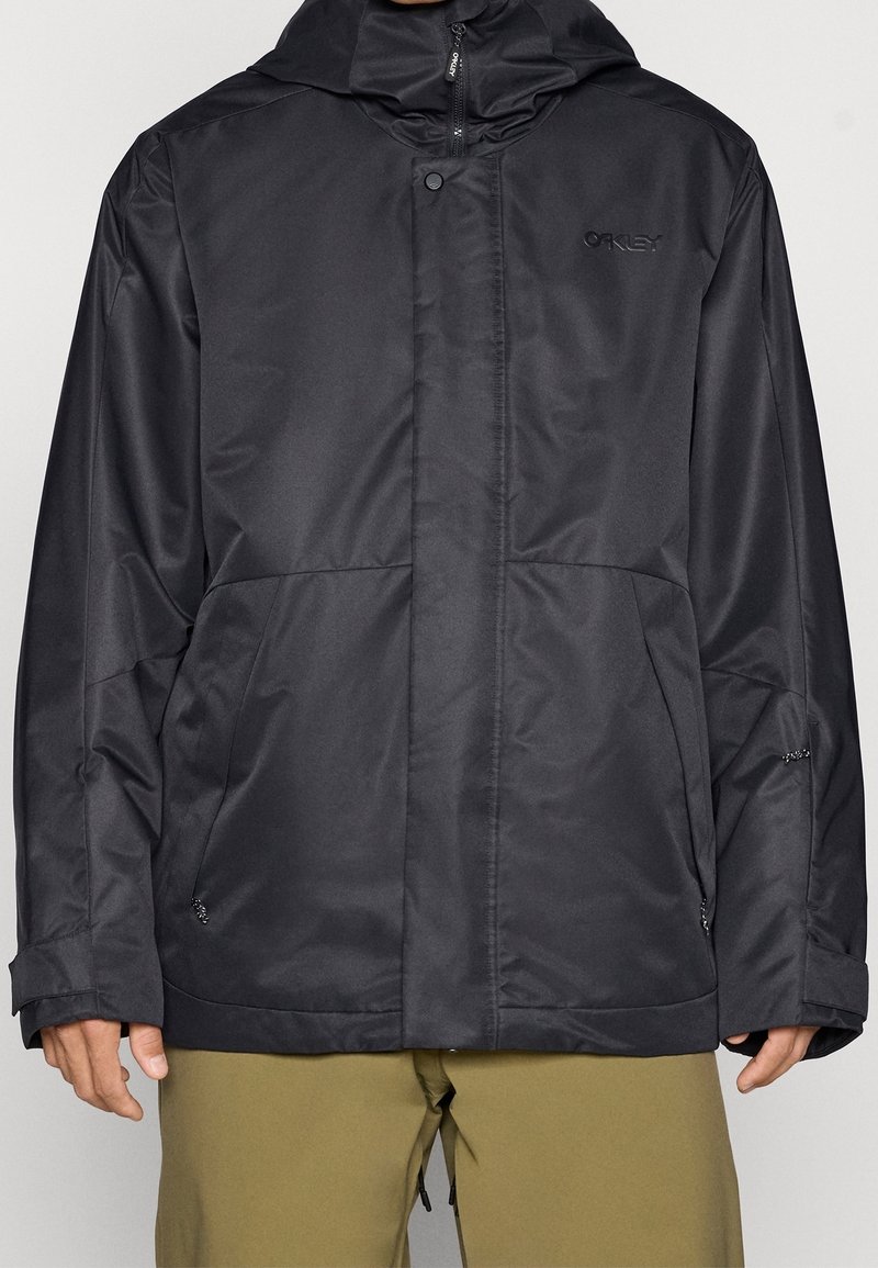 Veste à capuche noire Oakley avec fermeture éclair frontale et bouton-pression, portée par une personne avec un pantalon vert olive, mains visibles sur les côtés.