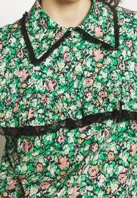 Blouse verte à fleurs avec des fleurs roses et blanches, bordure en dentelle noire sur le col et volant traversant la poitrine, boutons en perle sur le devant.