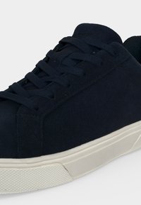 Gros plan d'une basket en daim bleu foncé avec des lacets assortis et une épaisse semelle en caoutchouc blanc cassé sur un fond clair.