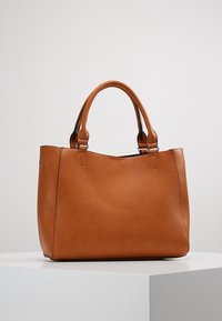 Brązowa skórzana torebka z podwójnymi okrągłymi uchwytami, prostokątnym kształtem i gładkim wykończeniem, eksponowana na białym piedestale na szarym tle.