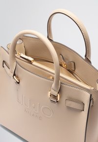 Sac à main beige en matériau synthétique texturé, avec des poignées doubles, des accents dorés, une marque en relief et un compartiment intérieur zippé.