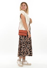 Vrouw in een wit mouwloos topje, leopardprintrok, beige sneakers, beige sokken en een roestkleurige crossbodytas die glimlacht terwijl ze opzij kijkt.