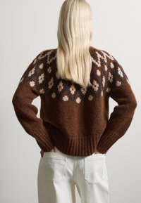 Suéter de punto marrón con un diseño floral en crema y azul, con dobladillo acanalado y ajuste holgado, llevado sobre pantalones anchos blancos.