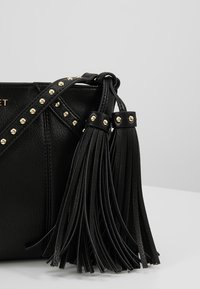 Borsa nera in pelle con borchie dorate, una lunga tracolla e dettagli in frange. La texture è liscia e presenta una forma strutturata.