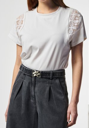 Vrouw met een wit t-shirt met kanten mouwdetails, ingestopt in hoge zwarte plissébroek met een zwarte, getextureerde riem.