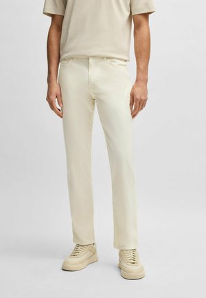 Mężczyzna w jasnych, dopasowanych spodniach w kolorze kremowym, beżowych trampkach oraz beżowej koszuli z krótkim rękawem, stojący na jednolitym jasnym tle.