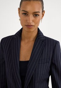 Moteris su atgal sušukuotais plaukais, dėvinti auksinius žiedinius auskarus ir tamsiai mėlyną dryžuotą švarką ant juodos palaidinės, žvelgianti į priekį su neutralia išraiška.