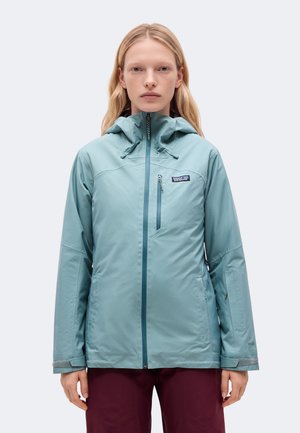 Une femme porte une veste Patagonia bleu clair à capuche, avec une fermeture éclair à l’avant et une poche poitrine, sur un fond uni.