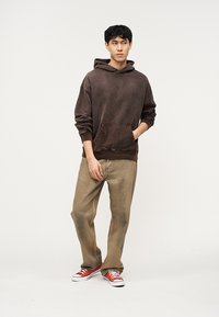 Νέος άνδρας που φορά καφέ φούτερ με κουκούλα, μπεζ παντελόνι και κόκκινα αθλητικά παπούτσια, στέκεται με το ένα χέρι στην τσέπη μπροστά σε απλό φόντο.