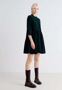 Robe en velours vert avec une fermeture à boutons à l'avant, des manches trois-quarts et une jupe évasée ; associée à des bottes en cuir marron jusqu'aux genoux.
