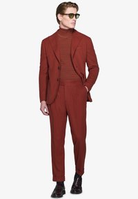 Rustfarvet jakkesæt lavet af tekstureret stof, med en tætsiddende jakke, matchende bukser og en ribbet turtle neck. Brune sko og solbriller.