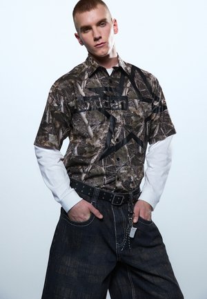 Ung mann med kort hår iført lagvis kamuflasje kortermet skjorte over hvit langermet skjorte, mørke vide jeans, og hendene i lommene.