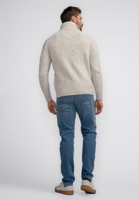 Pull en tricot beige avec un col montant, présentant un motif texturé. Associé à un jean bleu et des baskets grises et blanches.