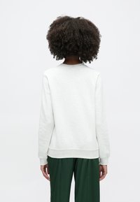 Άτομο με σγουρά μαλλιά που φοράει ανοιχτό γκρι μακρυμάνικο φούτερ και σκούρο πράσινο παντελόνι, στέκεται με την πλάτη γυρισμένη σε λευκό φόντο.