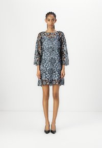 Spetsklänning med blå blommönster, trekvartsärmar, scallopad fåll, tillsammans med svarta högklackade skor, modell som står mot vit bakgrund.