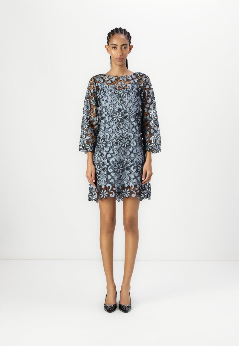 Spetsklänning med blå blommönster, trekvartsärmar, scallopad fåll, tillsammans med svarta högklackade skor, modell som står mot vit bakgrund.