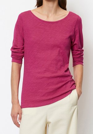 Woman wearing a long-sleeve dark pink top with ruched sleeves and off-white ribbed pants, standing with one hand in pocket.