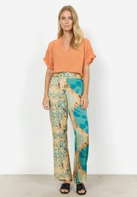 Orange Bluse mit kurzen Ärmeln und V-Ausschnitt; weit geschnittene Hose mit einem multicolor abstrakten Muster und Struktur; schwarze Sandalen.