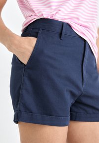 Person som bär marinblå shorts med ena handen i fickan och en rosa och vit randig skjorta som är lite nedstoppad.