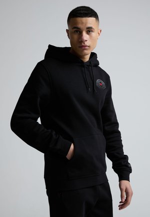 Ung mann iført en svart hettegenser med en logo på brystet, stående mot en ensfarget grå bakgrunn, med en hånd i lommen.