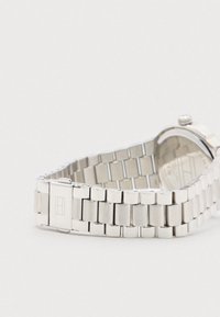 Ασημί μεταλλική μπάντα ρολογιού με κούμπωμα και μερικώς ορατό στρογγυλό καντράν σε απλό λευκό φόντο.