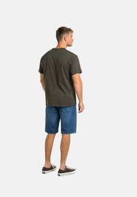 Camiseta de algodón verde oliva oscuro de manga corta, combinada con pantalones cortos de mezclilla azules y zapatillas marrones, vista desde la parte de atrás.