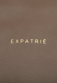 Barna bőr felület texturált befejezéssel. A "EXPATRIÉ" márkanevének arany betűkkel történő domborítása látható.