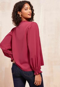 Blusa marrone con scollatura alta, maniche a sbuffo e polsini elastici; texture liscia, vestibilità leggermente ampia; abbinata a jeans in denim scuri.