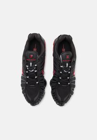 Schwarze Nike Shox Sneakers mit einem strukturierten Mesh-Obermaterial, roten Akzenten und charakteristischer gepolsterter Sohle. Mit weißen Schnürsenkeln und Markendetails.
